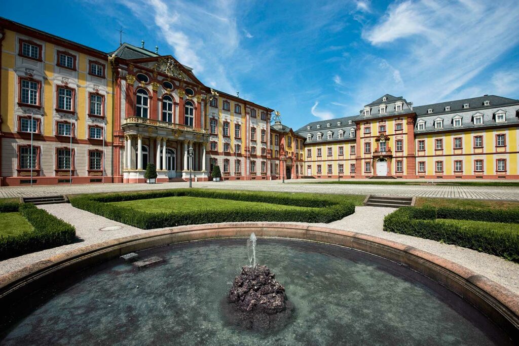 Foto vom Ehrenhof in Schloss Bruchsal, im Hintergrund zwei Flügel des Schlosses, im Vordergrund ein Springbrunnen mit Wasserbecken