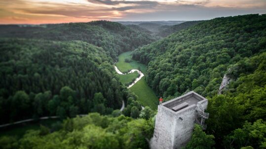 Foto vom Großen Lautertal aus der Vogelperspektive. Man sieht auch die Burgruine Wartstein, auf der ein Wanderer ins Tal blickt.