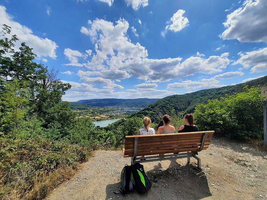 Foto von drei Frauen, die mit dem Rücken zur Kamera auf einer Bank sitzen und die Aussicht auf den Rhein genießen