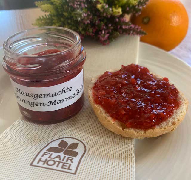 Foto von einem geöffneten Glas mit hausgemachter Blutorangenmarmelade und einer mit Blutorangenmarmelade bestrichenen Brötchenhälfte