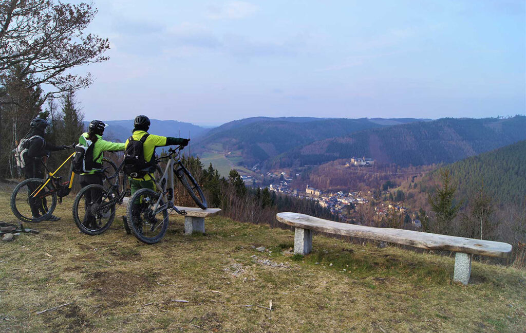 Foto von drei Mountainbikern, die an einem Rastplatz auf dem Berg ins Tal hinunterblicken.