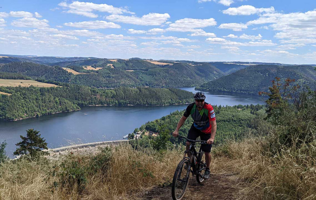 Foto von einem Mountainbiker, der hoch oben auf einem Berg am Rande eines Sees entlangfährt.